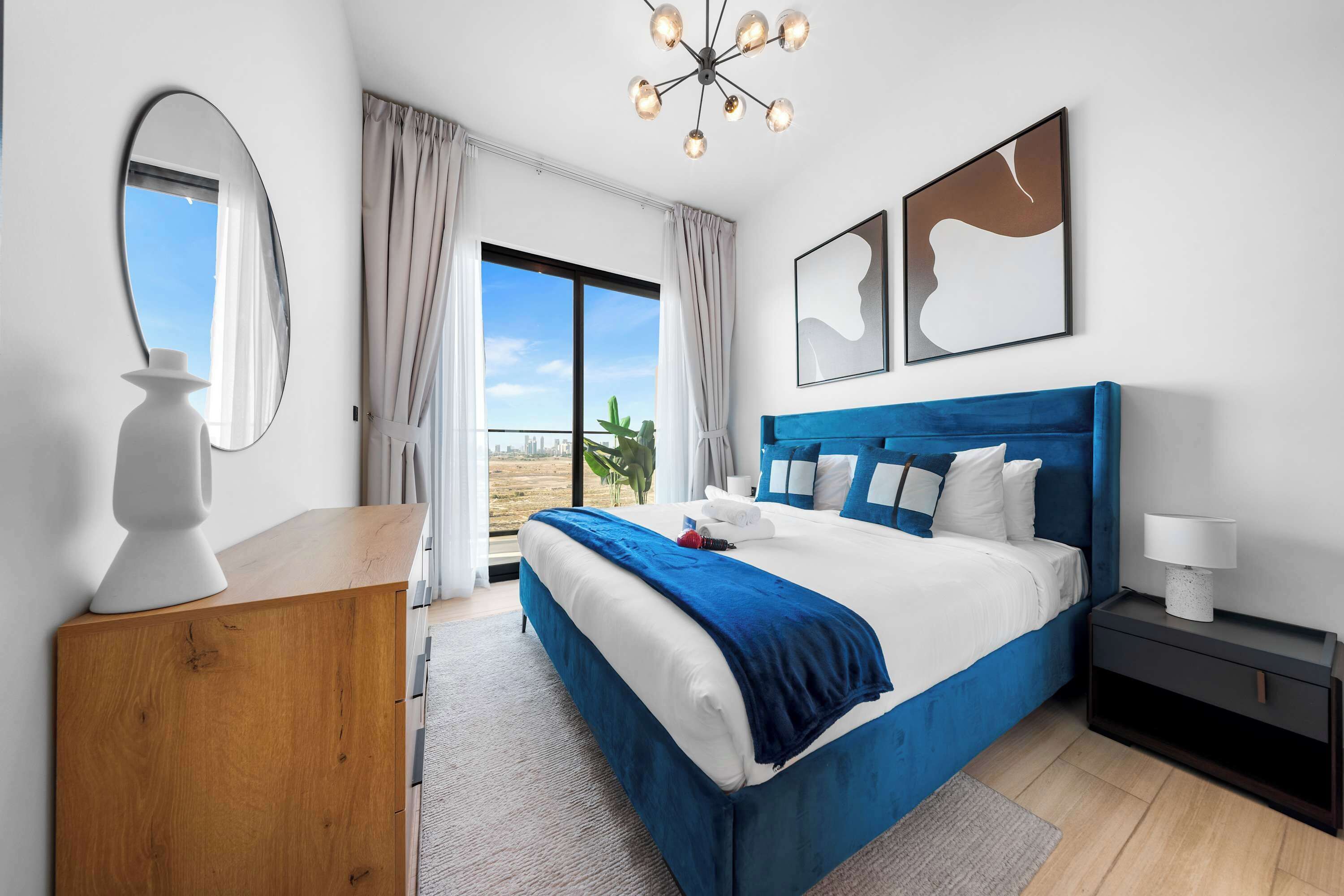 A modern bedroom with a large bed, a wooden dresser, a nightstand, and a view of the outdoors.