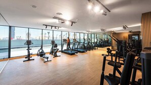 A modern gym with cardio equipment, weight machines, and a large window overlooking a cityscape. - StayShort - 1BR in JVC that Sleeps 4 with a Balcony! (JVC)