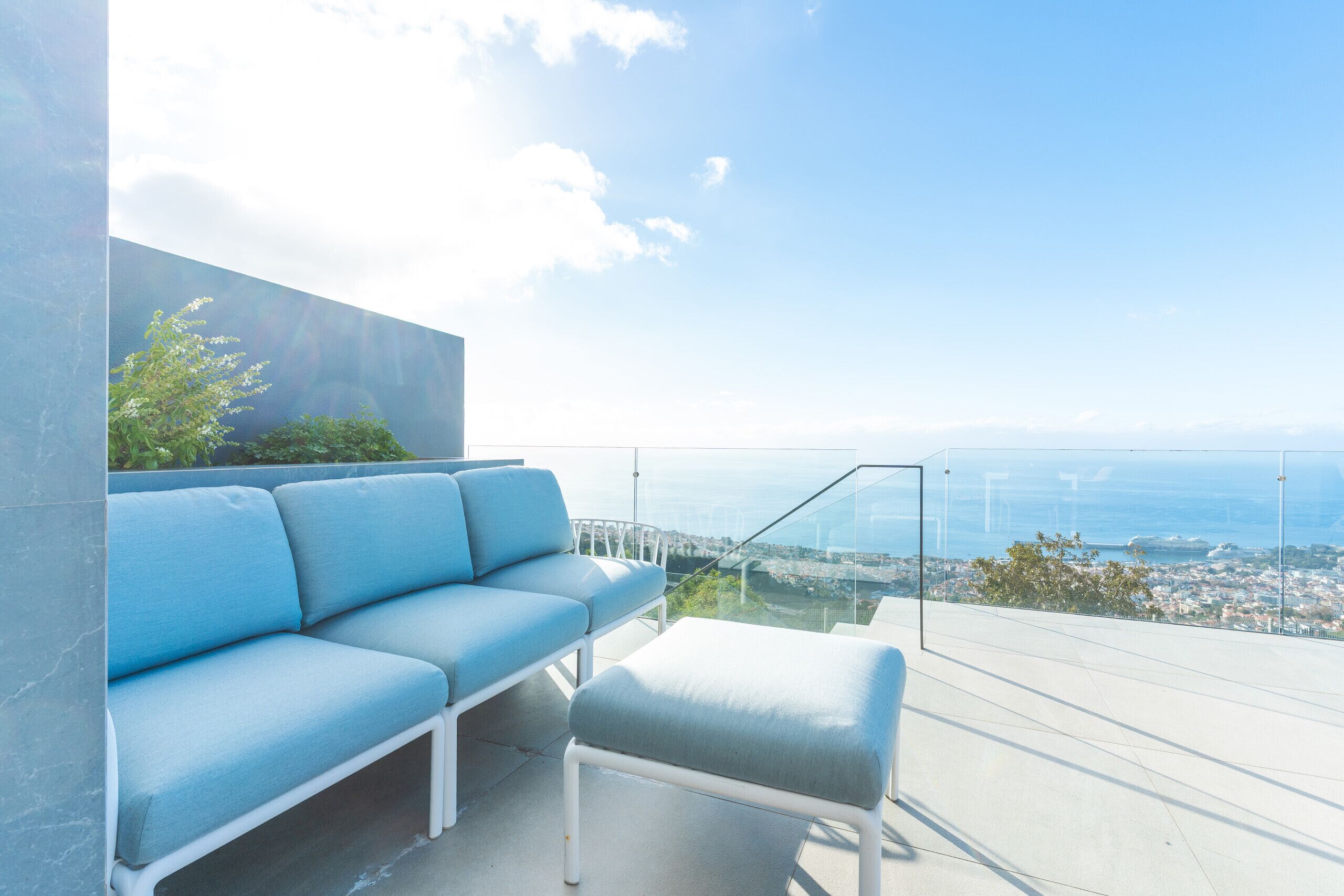 A modern outdoor seating area with a glass balustrade overlooking a cityscape and the sea.