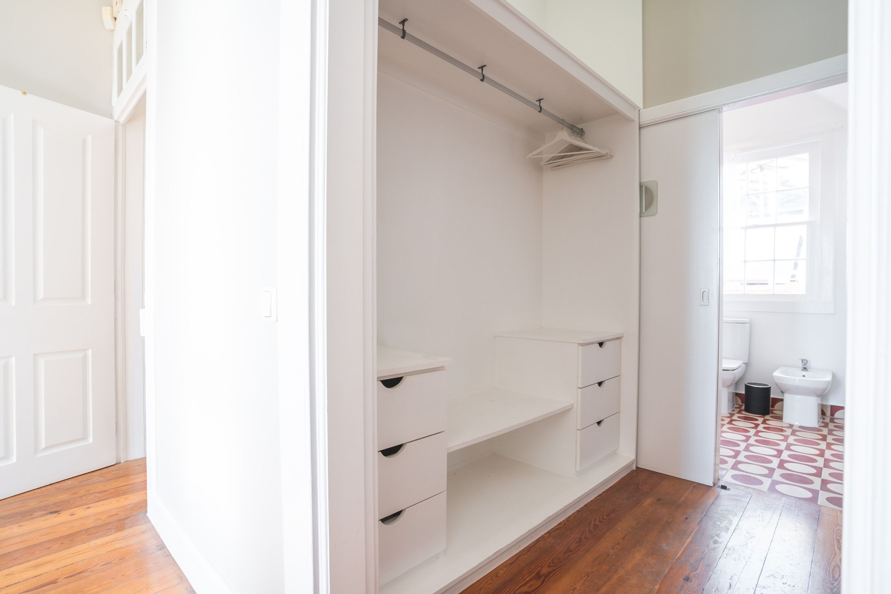 A room with a wooden floor, a white closet with hanging space, and a bathroom with a patterned floor.