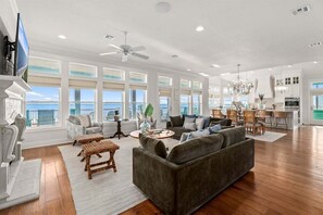 A spacious living room with a large gray sofa, a coffee table, and a TV. There are multiple windows offering a view of the ocean. - Beachfront | Swim Spa | Private Bowling Alley (Crystal Beach)