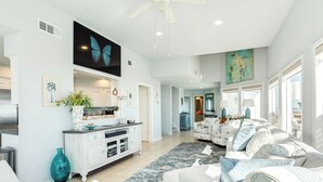 A spacious living room with a large sofa, a coffee table, and a dining area with a white cabinet and a large painting. - Beachfront Beauty w/ Wraparound Deck & Private Dune Walkover (Galveston)