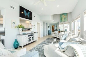 A spacious living room with a large sofa, a coffee table, and a dining area with a white cabinet and a large painting. - Beachfront Beauty w/ Wraparound Deck & Private Dune Walkover (Galveston)