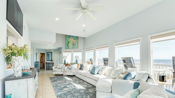 A spacious living room with a large window overlooking the ocean, a ceiling fan, and a modern white sofa set. - Beachfront Beauty w/ Wraparound Deck & Private Dune Walkover (Galveston)