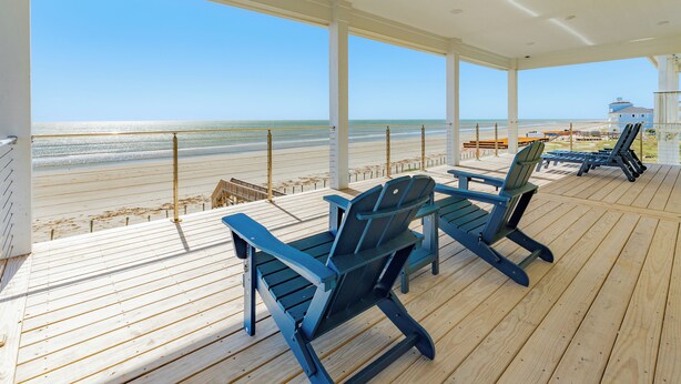 Une terrasse couverte avec un plancher en bois et des chaises Adirondack bleues, donnant sur une plage sous un ciel dégagé.