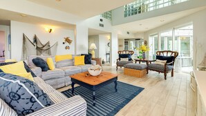Un salon spacieux avec un plafond haut, une grande fenêtre et une vue sur l’extérieur. On y trouve un canapé, une table basse et deux fauteuils.