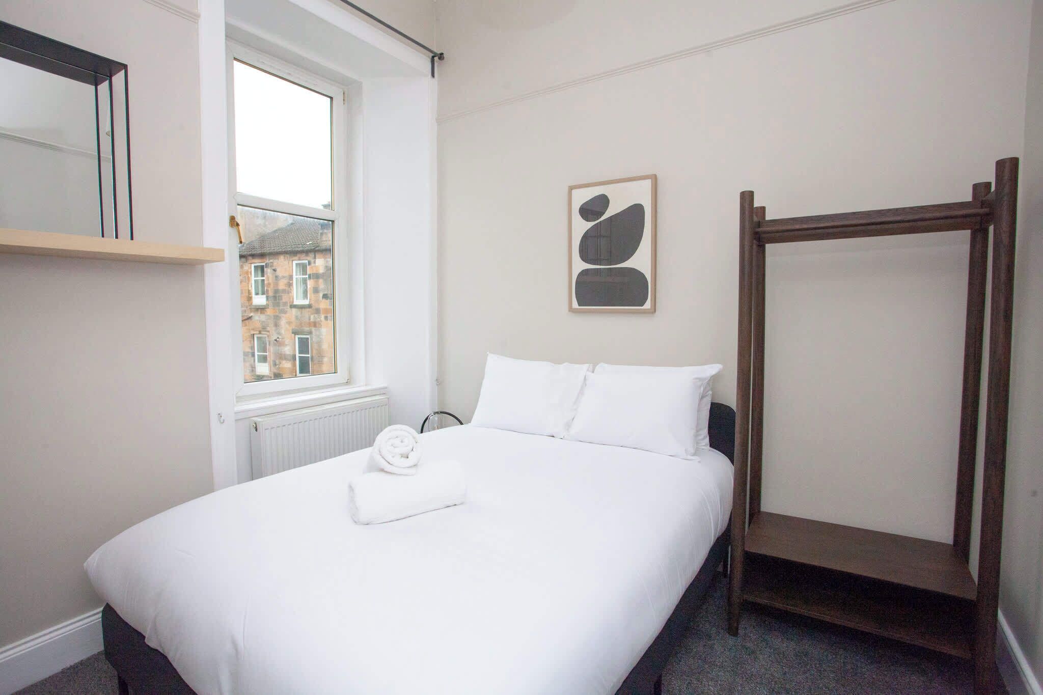 A hotel room with a bed, a wooden bench, a window, and a framed artwork on the wall.