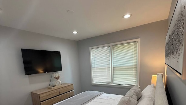 A modern bedroom with a large bed, a TV mounted on the wall, a window with blinds, and a bedside table with a lamp. - Ranch Cozy Home close to O'Hare (River Grove)