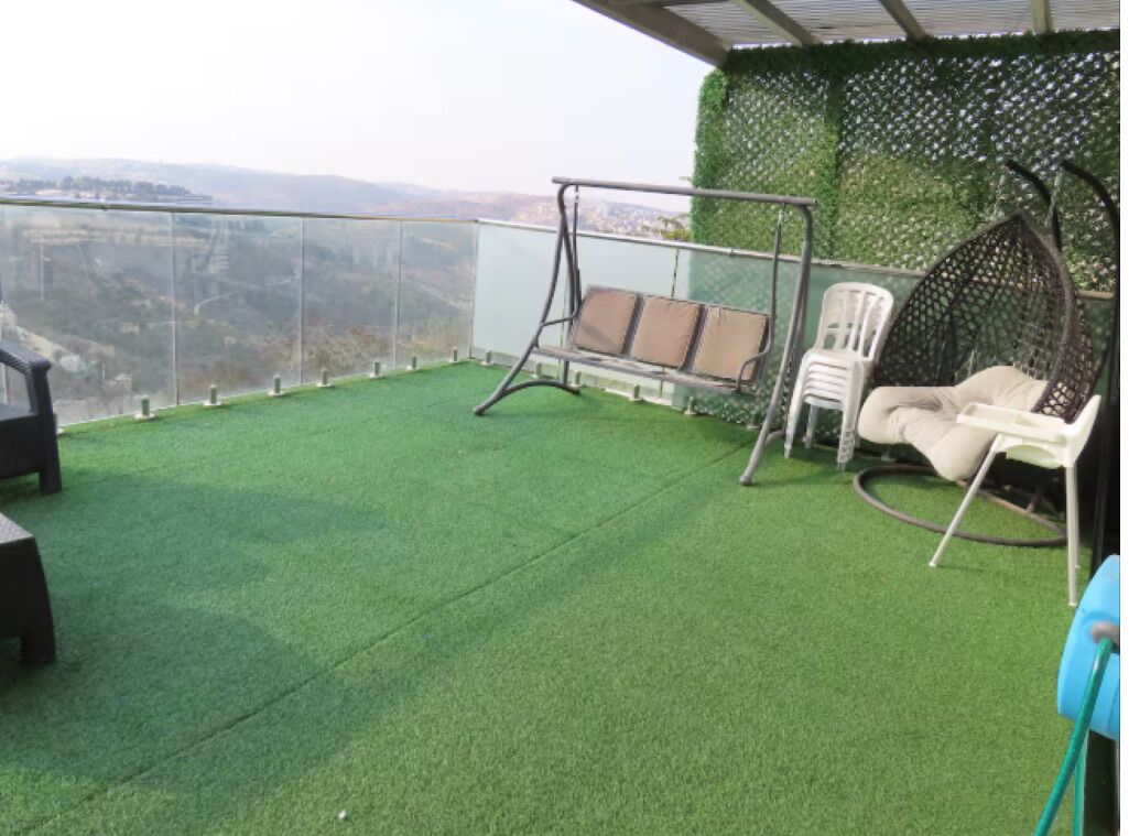 Un balcon agrémenté de gazon synthétique, d’une chaise suspendue, d’un fauteuil en osier et offrant une vue sur la campagne.