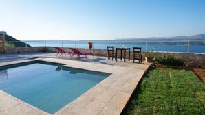 A swimming pool with lounge chairs and a table, surrounded by a stone wall and a grassy area, with a view of the sea and distant mountains. - Aptera Dream Apartment (Souda)