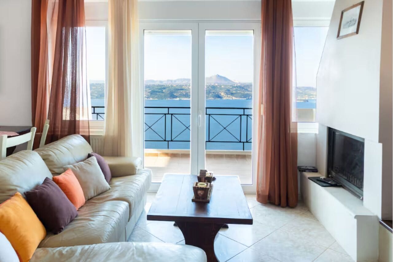 Ein Wohnzimmer mit einer Couch, einem Couchtisch und einem Fernseher. Es gibt einen Balkon mit Blick auf das Meer und die Berge.
