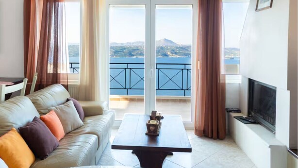 A living room with a sofa, a coffee table, and a TV. There is a balcony with a view of the sea and mountains. - Aptera Dream Apartment (Souda)