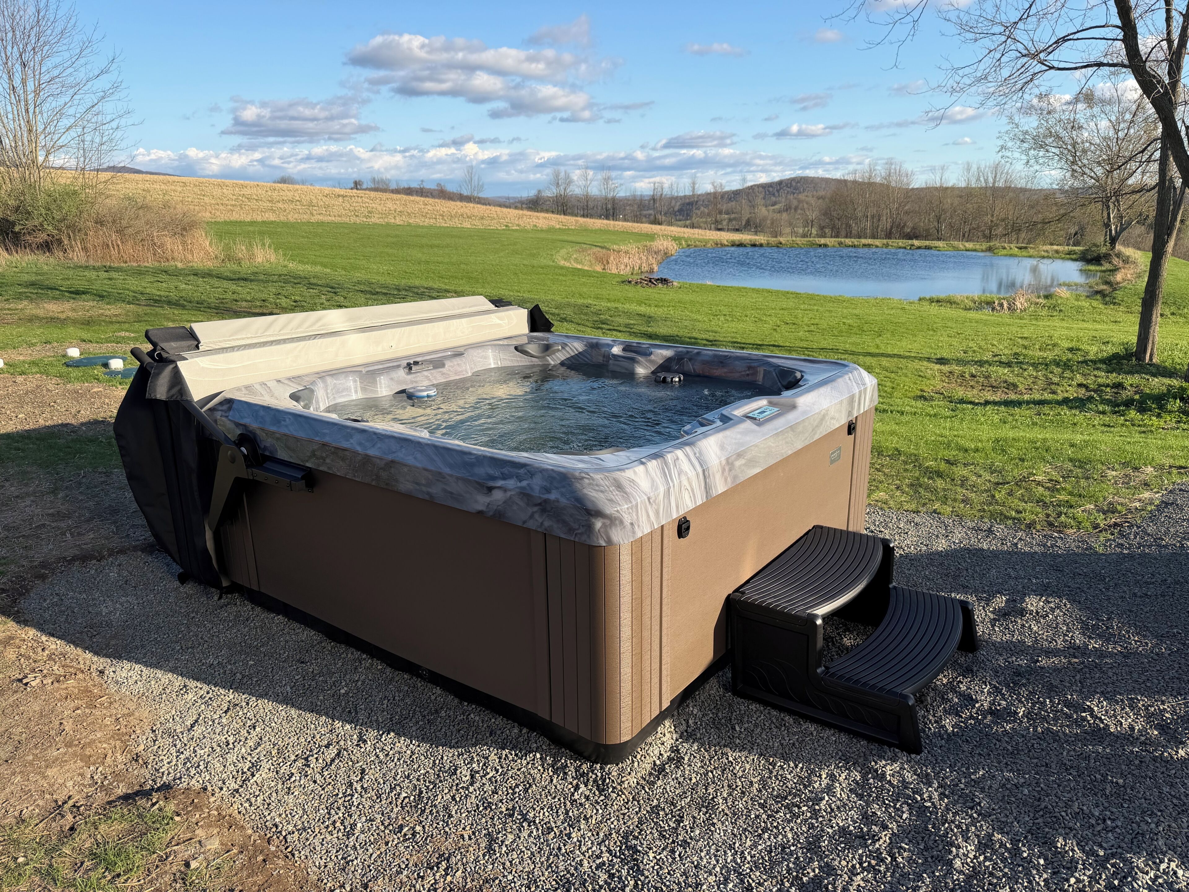 Un bain à remous extérieur, installé sur une base de gravier, entouré d’un champ herbeux et d’un lac.