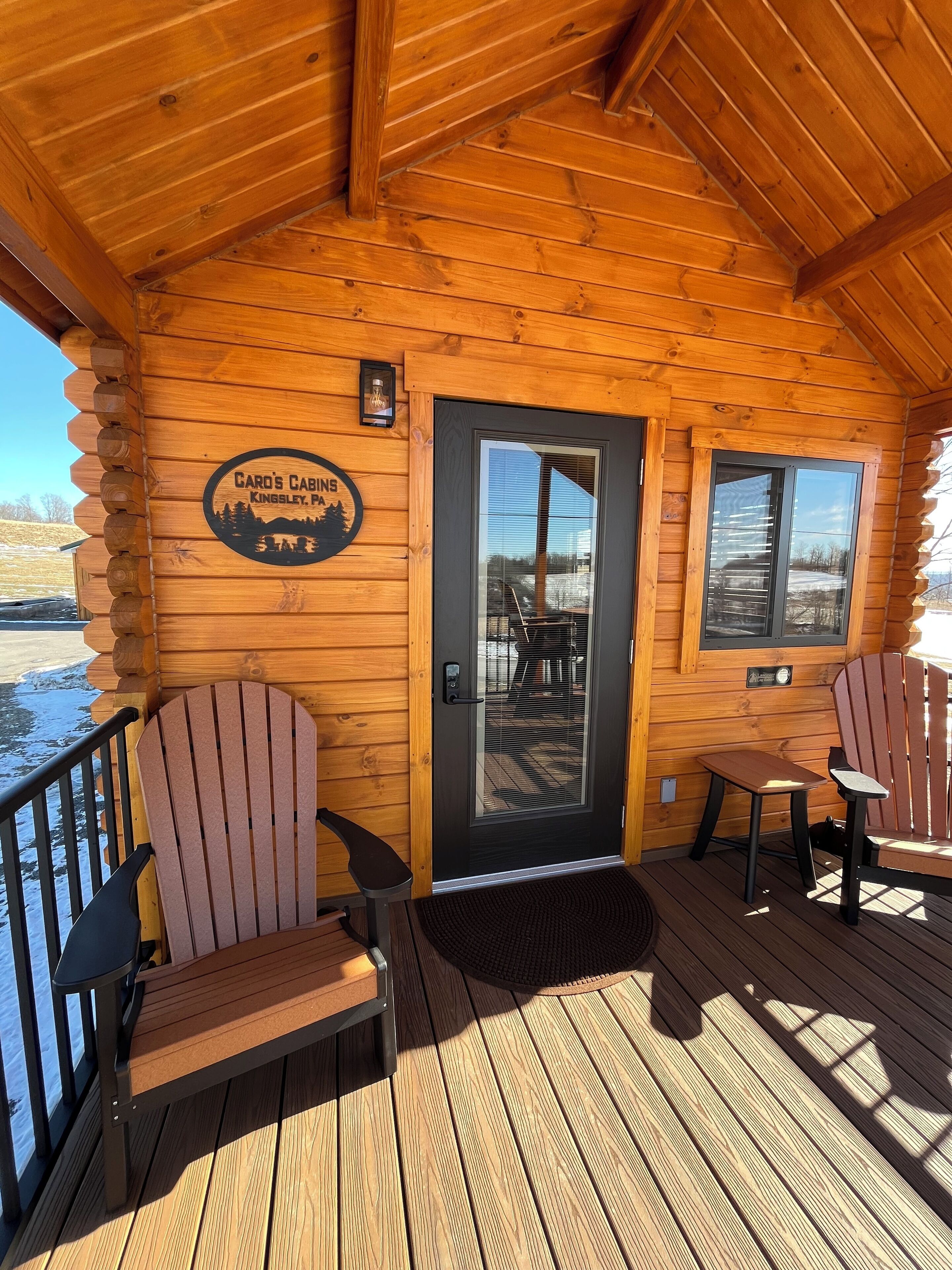 A log cabin with a wooden deck, two Adirondack chairs, a table, and a door with a sign reading "Card's Cabs Kingsley, PA".