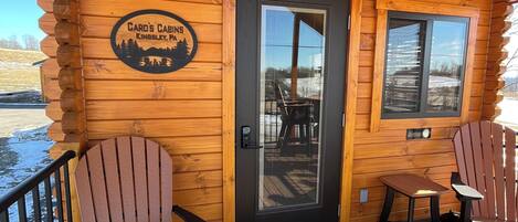 A log cabin with a wooden deck, two Adirondack chairs, a table, and a door with a sign reading "Card's Cabs Kingsley, PA".