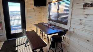 Interior d'una cabana de fusta amb taula de dinar, cadires, una banca, un televisor muntat a la paret i una finestra amb persianes.