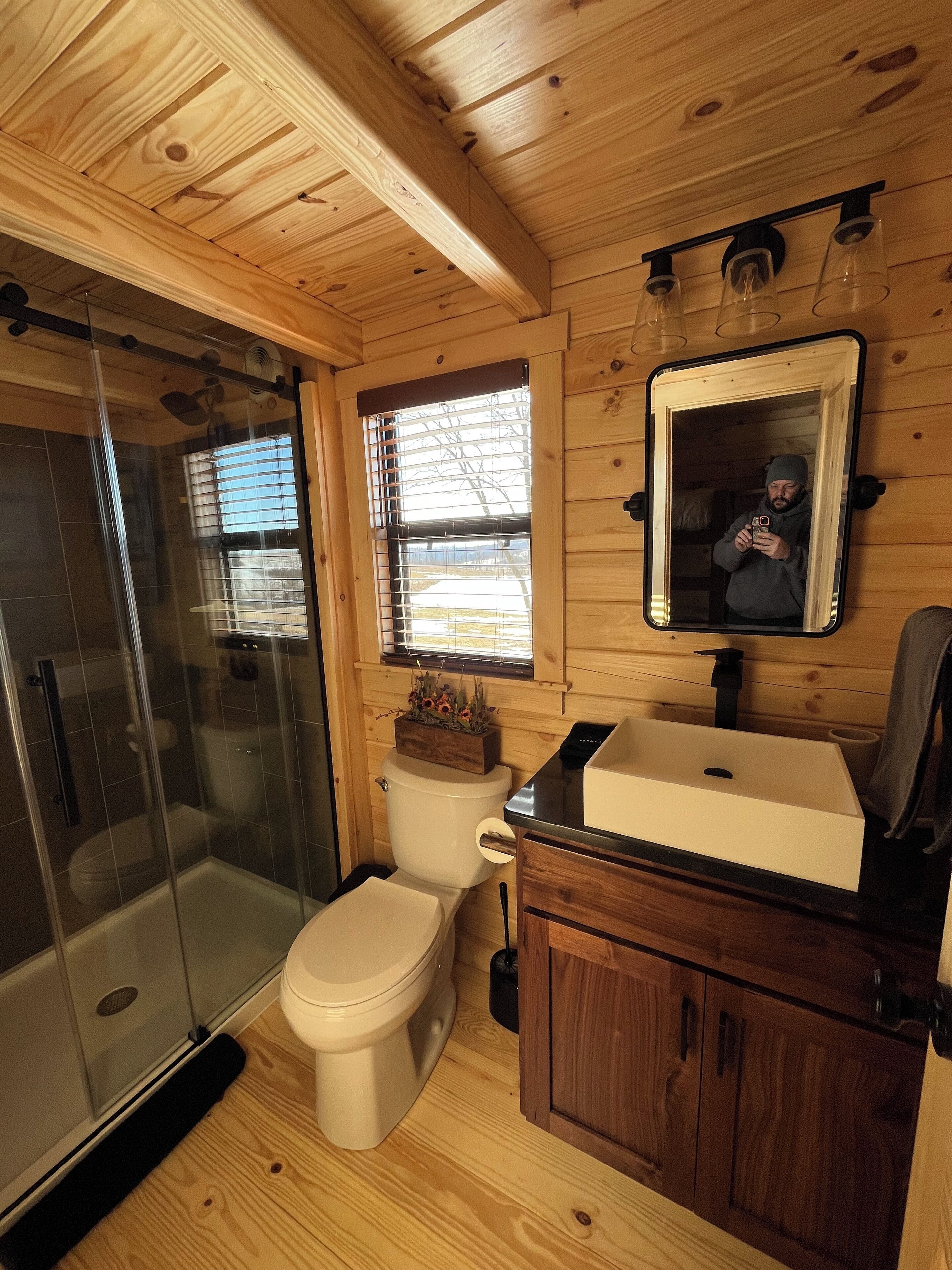 Un baño de cabaña de madera con mampara de vidrio, inodoro, lavamanos y espejo.