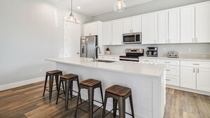 Une cuisine moderne avec des meubles blancs, des appareils en acier inoxydable, un grand îlot central doté de tabourets et un éclairage suspendu.