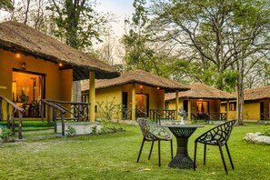 A bungalow-style accommodation with thatched roofs, a small outdoor seating area, and a well-maintained lawn. - Bijrani Jungle Cottages by CYJ (Ramnagar)