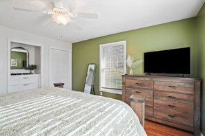 Une chambre à coucher comprenant un lit, une commode, un téléviseur, un ventilateur de plafond et une fenêtre avec des stores.