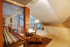 A modern loft bedroom with a large window, a bed with patterned bedding, a chair, and a floor lamp.
