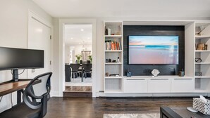 En moderne stue med et fladskærms-tv monteret på et hvidt tv-møbel, et spiseområde med et bord og stole i mørkt træ, og en gang, der fører til et andet rum.