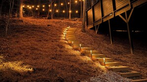 Ein hölzerner Weg mit Lichterketten führt zu einer überdachten Holzkonstruktion in bewaldetem Gelände.