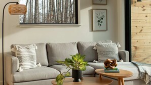 Ein modernes Wohnzimmer mit einem grauen Sofa, einem runden Holztisch und einem großen Fenster mit Blick auf Bäume.