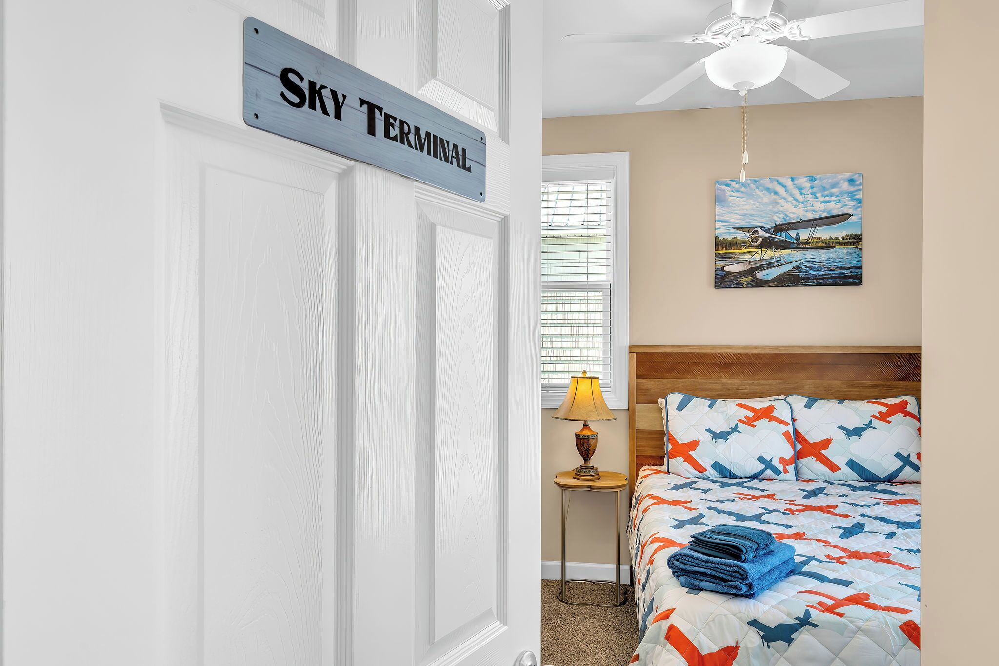 A bedroom with a bed, a nightstand, a lamp, a ceiling fan, a painting, and a sign that reads "SKY TERMINAL".