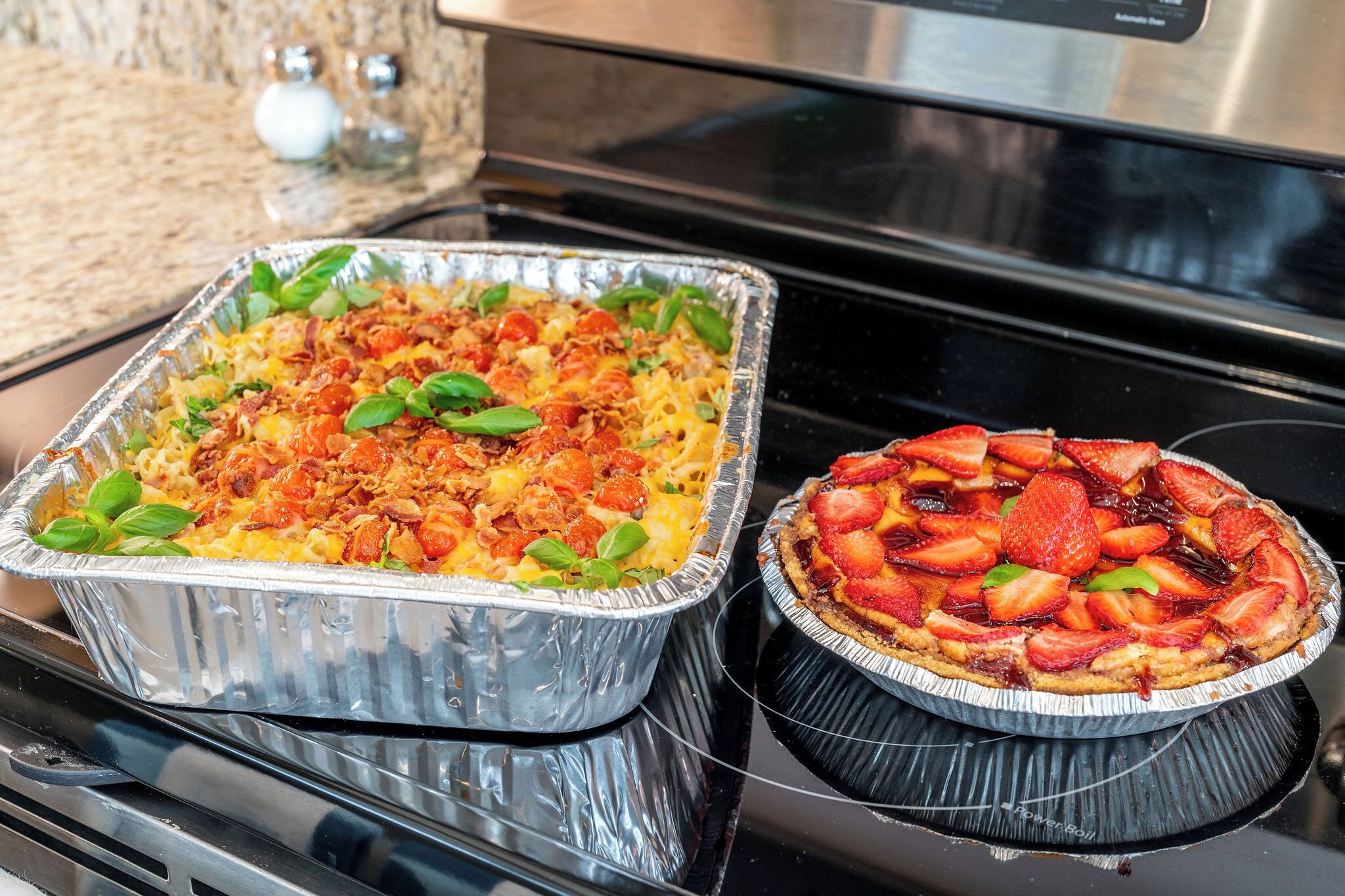 A kitchen with a rectangular aluminum tray containing a casserole dish and a round aluminum pie on a stovetop.