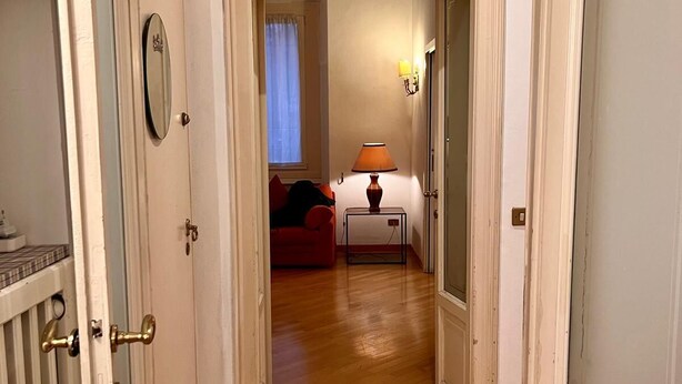 A hallway leading to a bedroom with a wooden floor, a red sofa, a bedside table with a lamp, and a window with curtains. - Elegant & Large flat for Family and Groups (Milano)