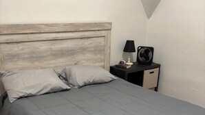 Un lit bien fait, avec une couette et des oreillers gris, une tête de lit en bois, une table de chevet avec une lampe et un ventilateur, et un petit tiroir.