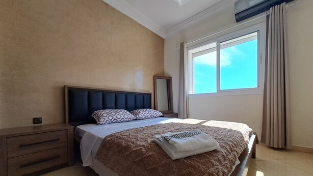 Un dormitorio con cama, mesita de noche, ventana con cortinas y aire acondicionado instalado en la pared.