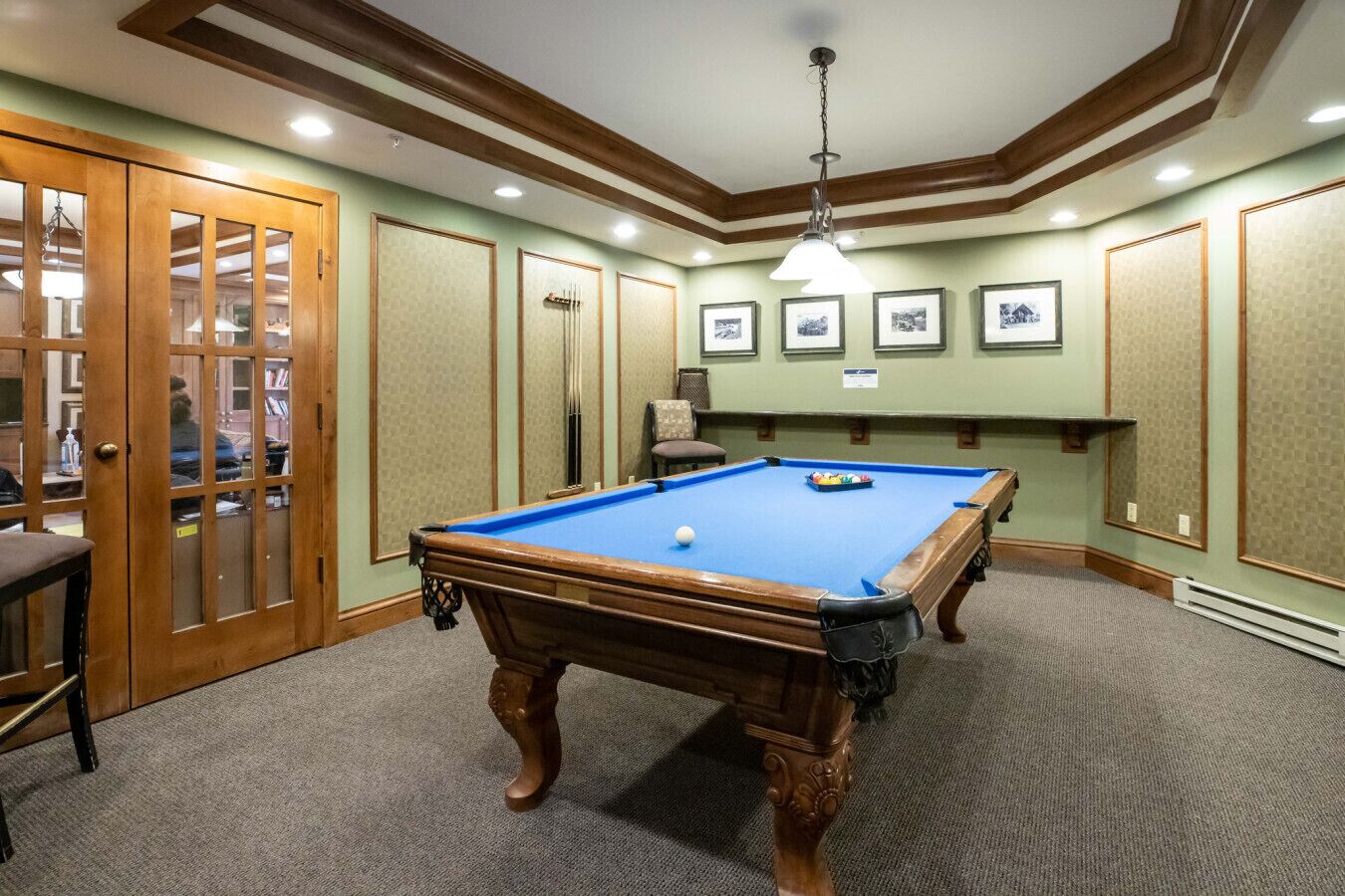 Mesa de bilhar num espaço com paredes verdes, painéis de madeira e quadros enquadrados.