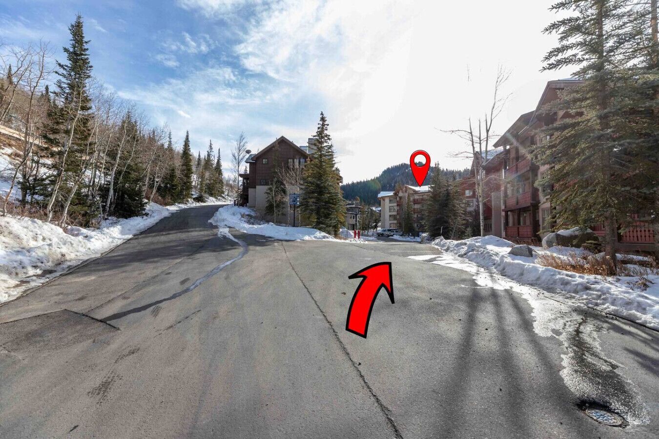 A snow-covered road leading to a building with a red roof, surrounded by trees and a mountainous backdrop.