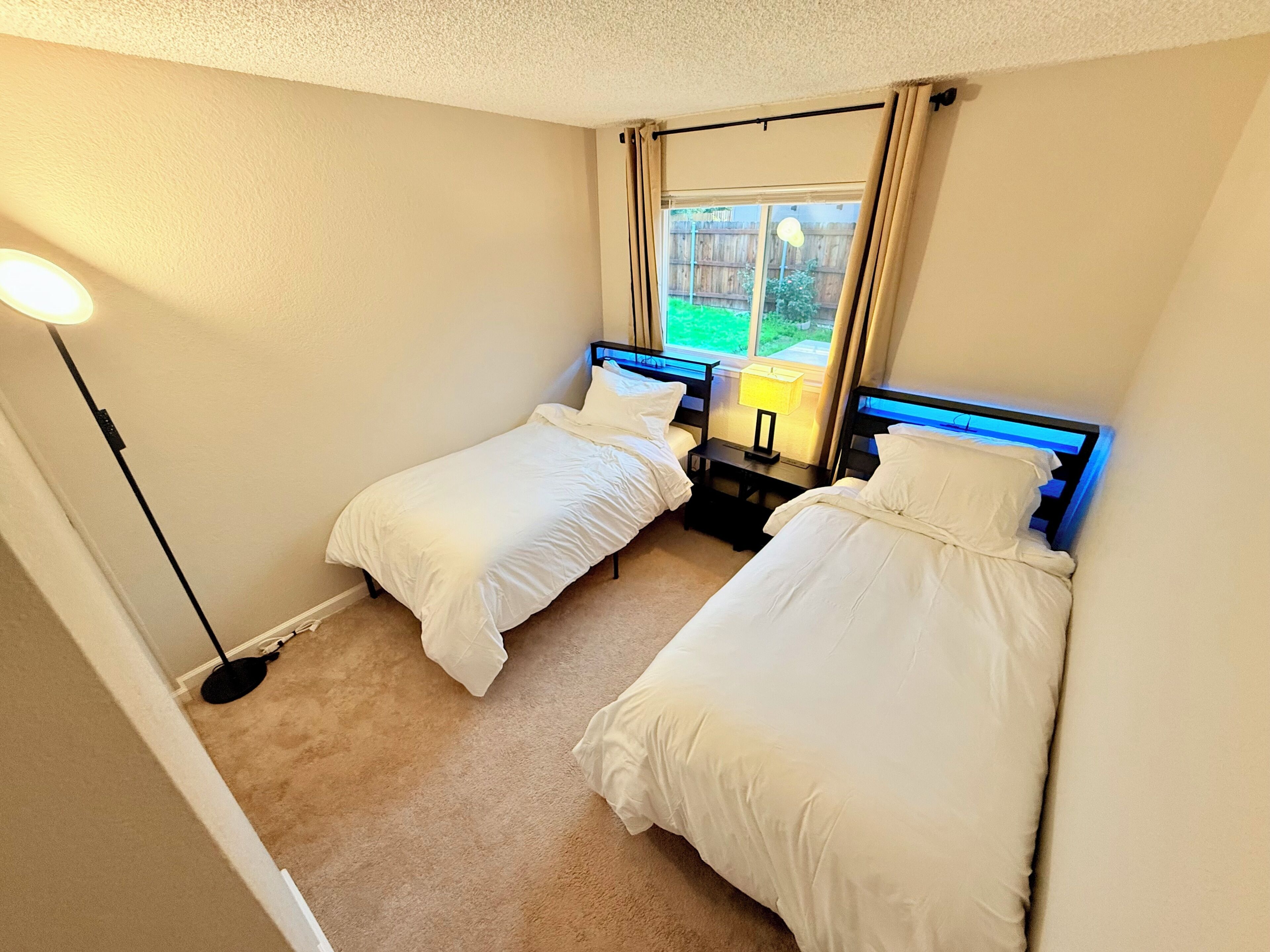A twin room with two beds, a window with curtains, a bedside table with a lamp, and a floor lamp.