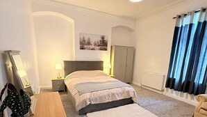 Une chambre à coucher comprenant un lit, des lampes de chevet, un bureau avec une chaise, un radiateur et une fenêtre avec des rideaux.