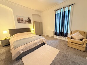 Un dormitorio con cama, una silla, una mesita de noche, lámpara, armario y ventana con cortinas.