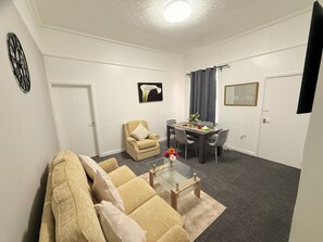 A living room with a sofa, a coffee table, a dining table with chairs, a small table with a vase, and a wall-mounted TV. - Cosy Two-bedroom Bungalow in the Centre of Sunderland (Sunderland)