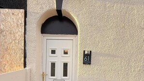 Une porte blanche surmontée d’une arche noire, une boîte aux lettres portant le numéro 61 et un coffret de raccordement électrique blanc.