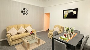 Un salon avec un canapé beige, une table basse en verre, une table à manger avec des chaises, une horloge et un tableau accroché au mur.