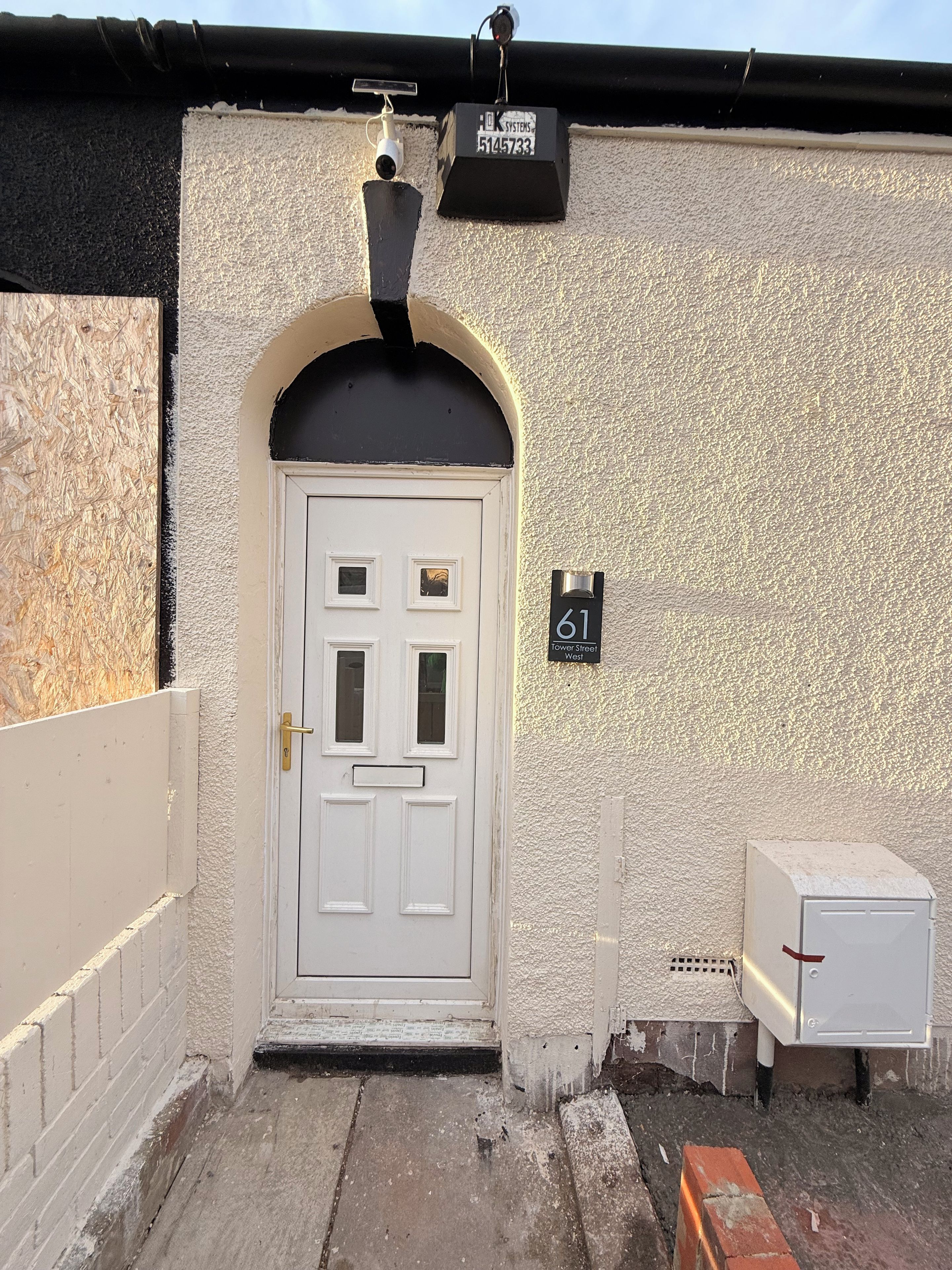 A white door with a black arch, a mailbox with the number 61, and a security camera on the wall.