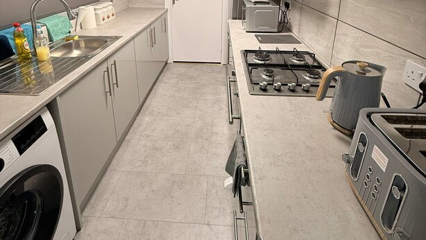 Une cuisine moderne équipée d’un four encastré, d’une cuisinière à gaz et d’un four à micro-ondes. On y trouve un évier avec un robinet, ainsi qu’une porte donnant sur une autre pièce.