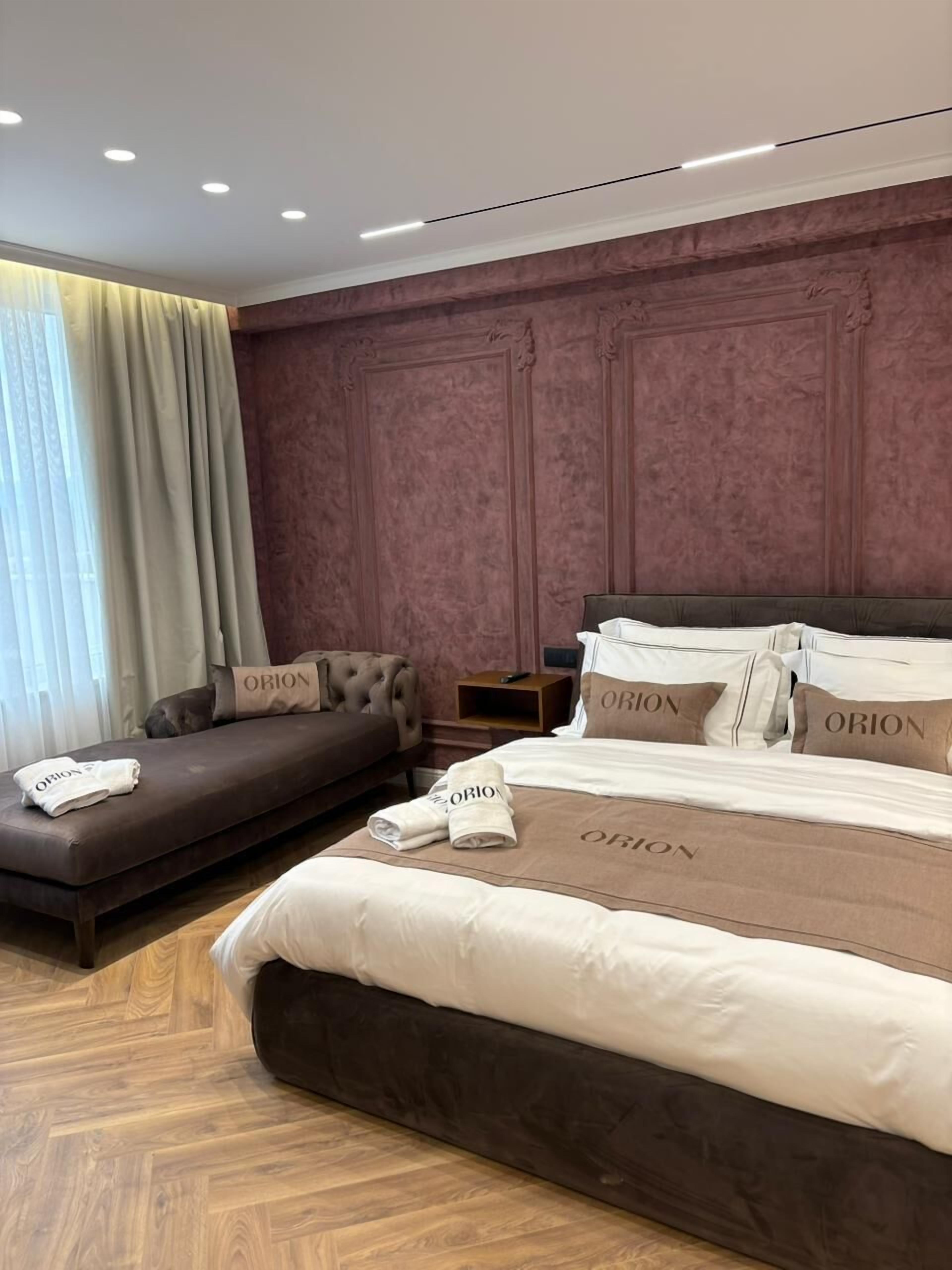 Ein Hotelzimmer mit einem großen Bett, einer Couch und einem Nachttisch. Das Bett hat mehrere Kissen mit dem Schriftzug "ORION". Der Raum hat einen Holzboden und ein großes Fenster mit Vorhängen.
