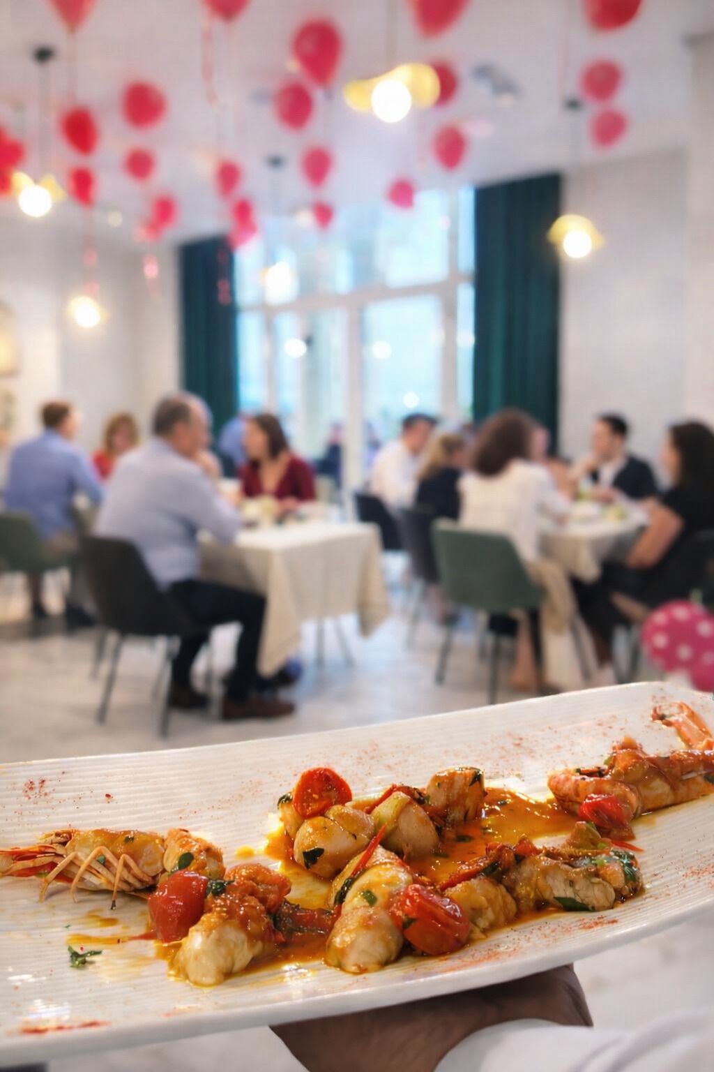 A dining area with tables and chairs, a plate of food in the foreground, and red balloons in the background.
