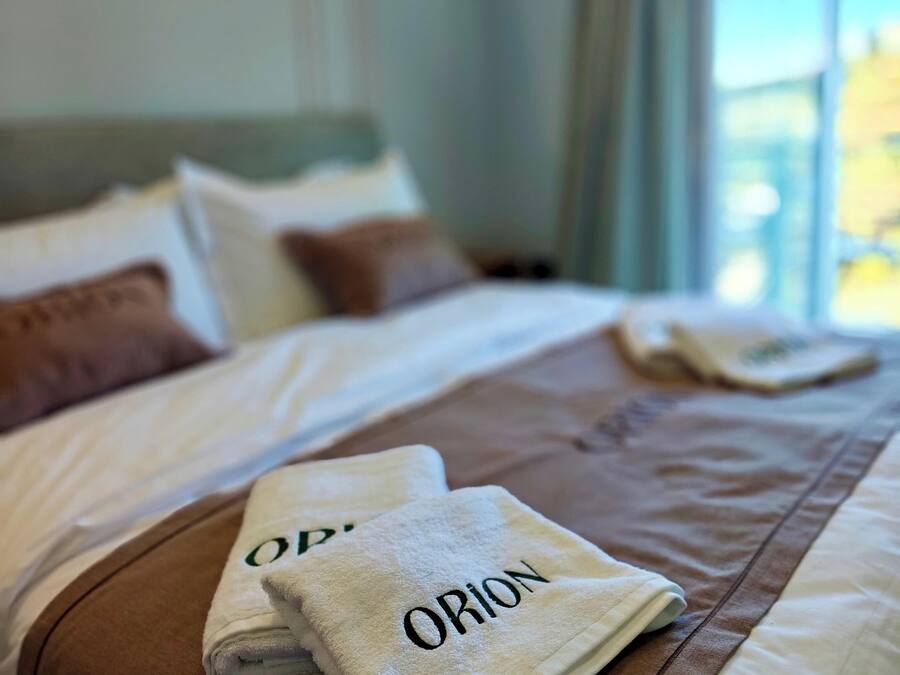 A neatly made bed with a white and brown bedspread, two folded towels with the word "ORION" on them, and a window with curtains in the background.