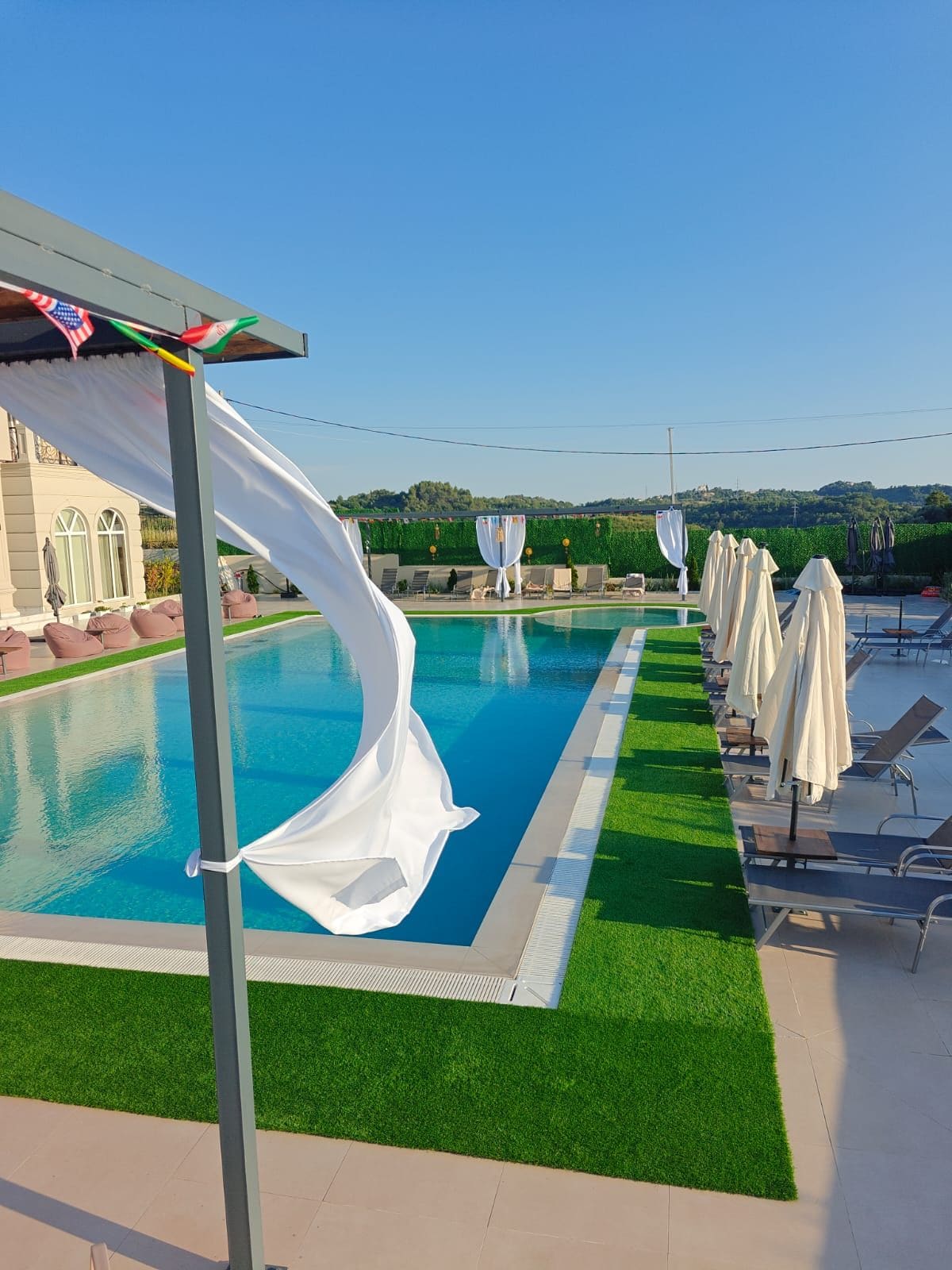 A swimming pool with a clear blue water, surrounded by artificial grass, and a row of sun loungers with white umbrellas.