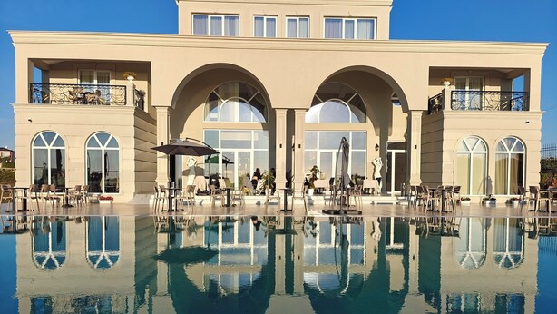 Un edificio grande con piscina que refleja su arquitectura, mobiliario exterior y cielo despejado.