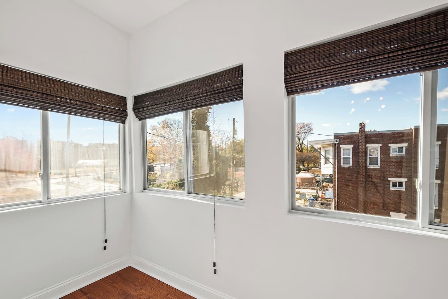 Una habitación con tres ventanas grandes, cada una con una persiana oscura, paredes blancas y suelo de madera.