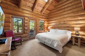 A log cabin bedroom with a large bed, two windows, a desk, and a chair.